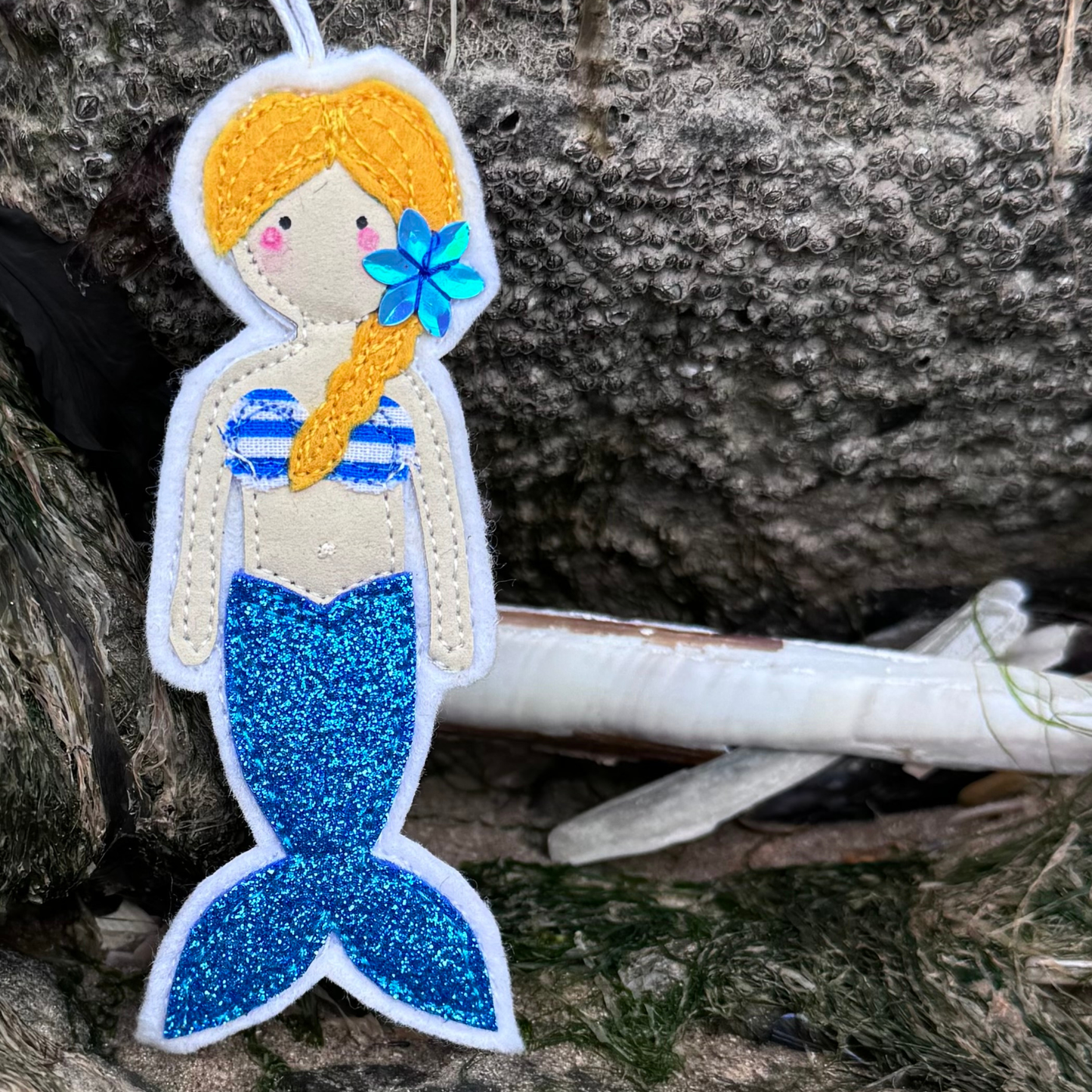 Handmade blue glitter tail mermaid hanger with striped bikini top displayed on coastal rocks. The perfect gift for wild swimmers and sea dippers, only from Waves & Wild Water.