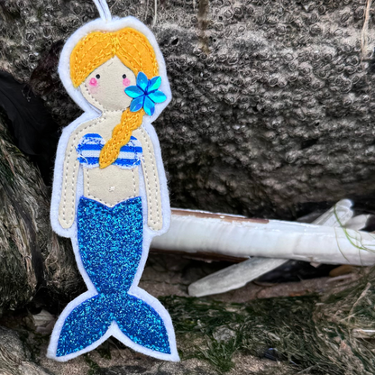 Handmade blue glitter tail mermaid hanger with striped bikini top displayed on coastal rocks. The perfect gift for wild swimmers and sea dippers, only from Waves & Wild Water.