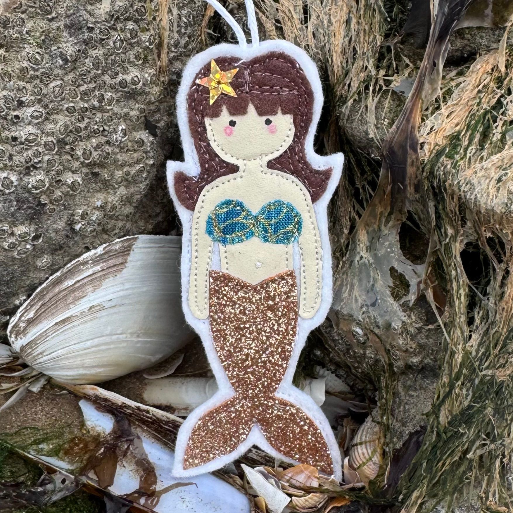 Handmade gold glitter tail mermaid hanger with patterned bikini top displayed on coastal rocks with shells. Only from Waves & Wild Water.