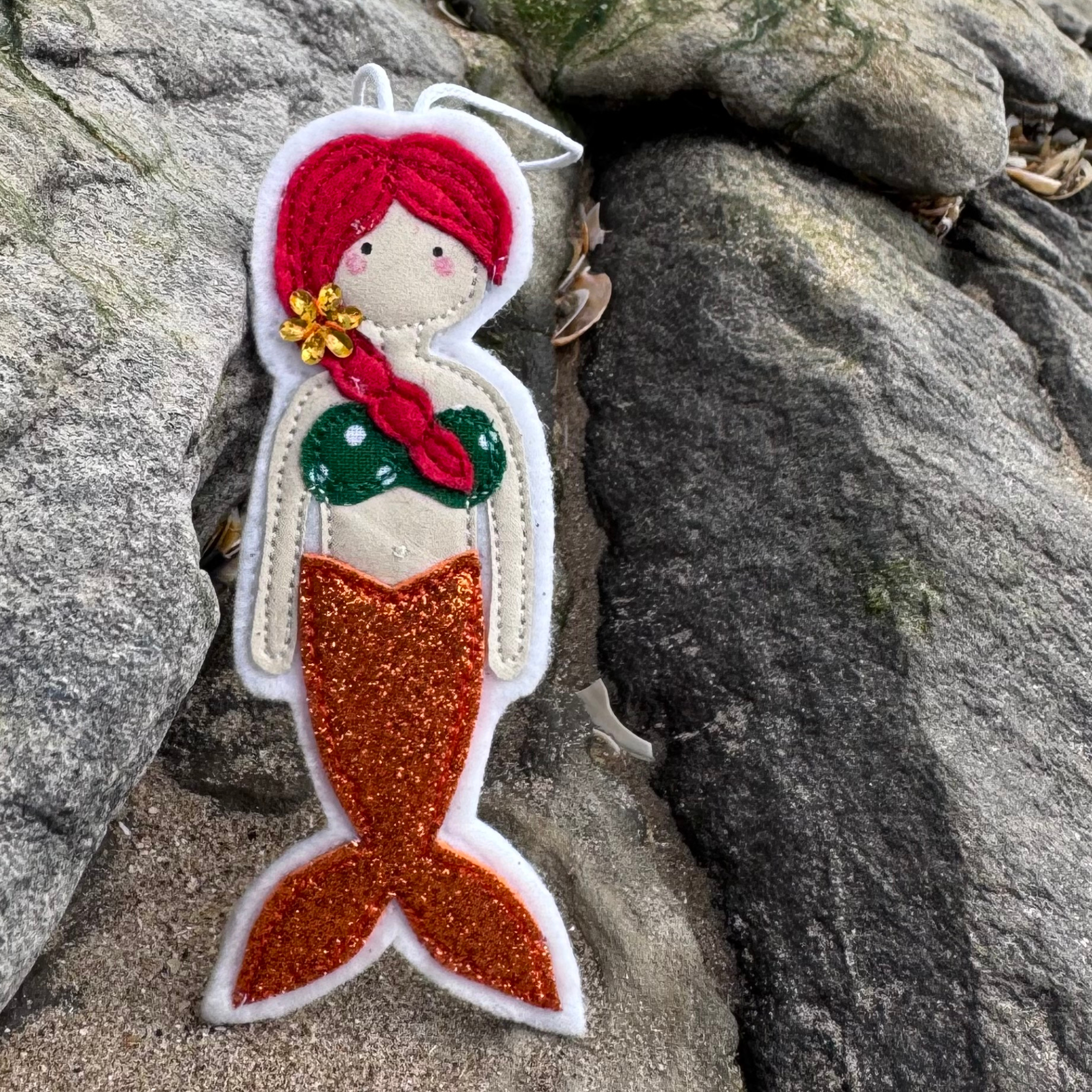 Handmade felt mermaid decoration with orange glitter tail and green polka dot top, displayed on coastal rocks. From the Wild Water Mermaids collection, only from Waves & Wild Water.