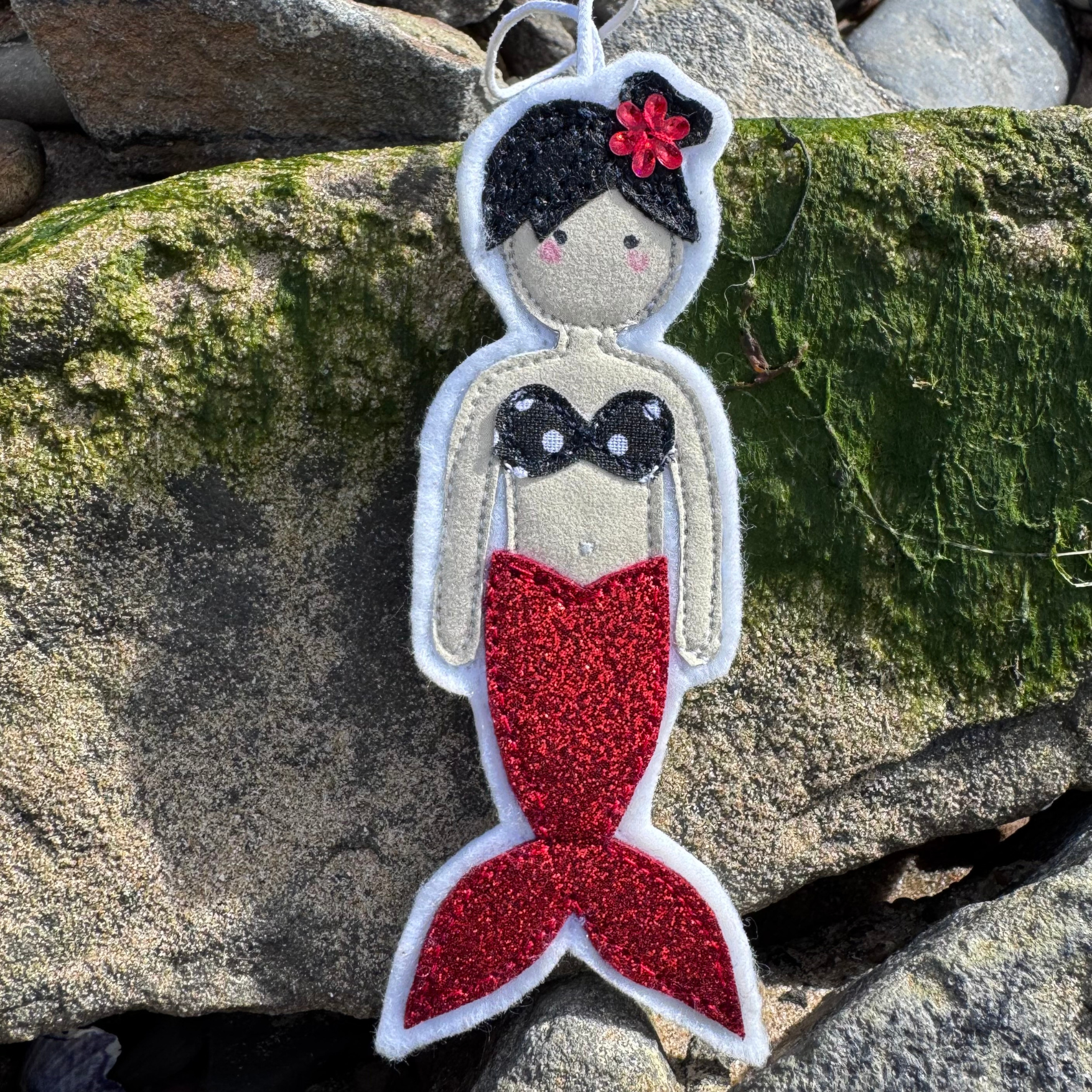 Handmade red glitter tail mermaid hanger with braided hair and black bikini top photographed on seaweed-covered rocks at the beach.