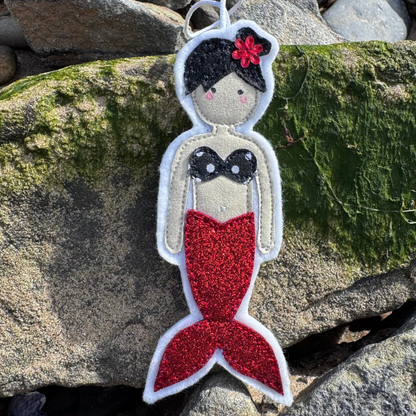 Handmade red glitter tail mermaid hanger with braided hair and black bikini top photographed on seaweed-covered rocks at the beach.