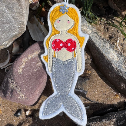 Handmade felt mermaid hanger with silver glitter tail and red polka dot top, displayed on sandy beach with shells and coastal stones. Only from Waves & Wild Water.