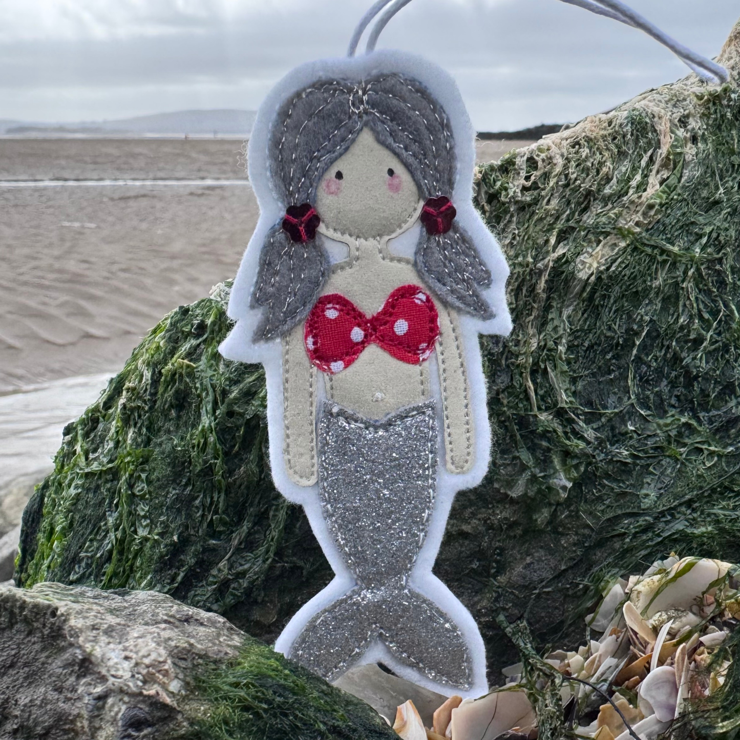 Handmade silver glitter tail mermaid hanger with red polka dot bikini top photographed on seaweed-covered rocks at the beach.