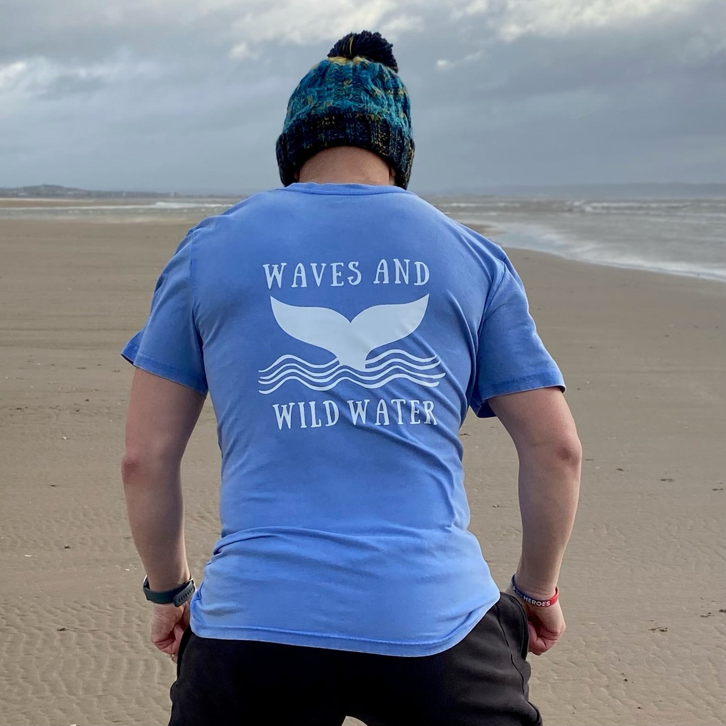 A man on the beach wearing a blue t-shirt with Waves and Wild Water printed on the back.