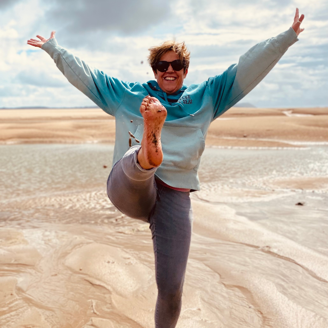 A woman wearing a blue hoodie on the beach with her arms in the air and kicking her foot up.