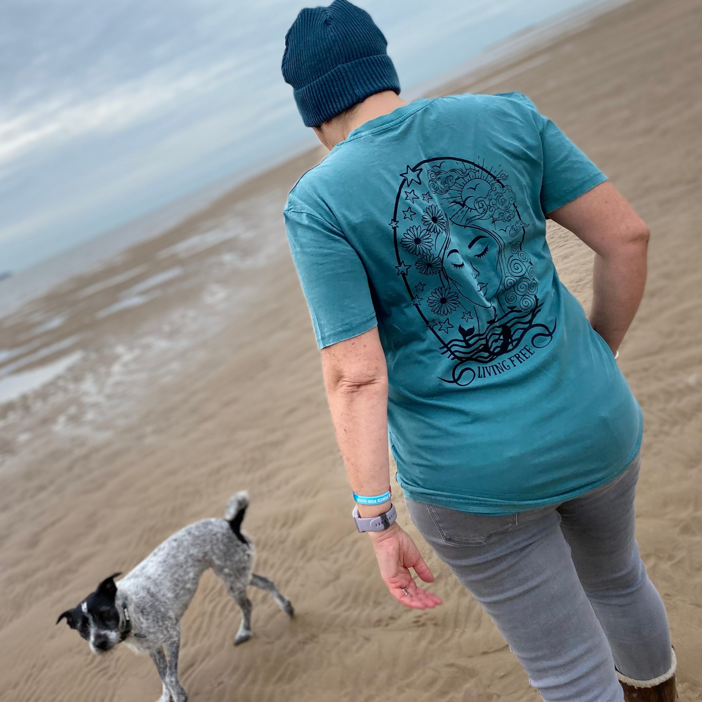 A woman in a t-shirt and hat, with a dog on the beach.