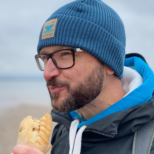 A man with a beard, wearing glasses and a teal green fisherman beanie hat, while eating a pasty.