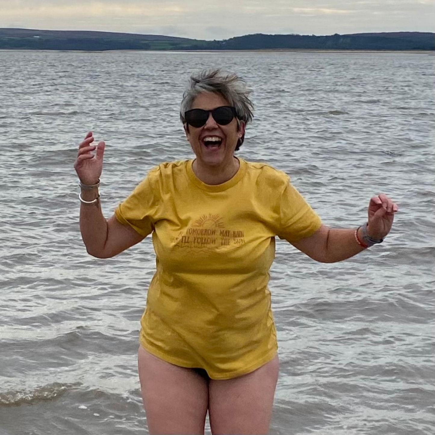 A woman laughing and dancing in the sea wearing a vintage gold t-shirt and sunglasses.