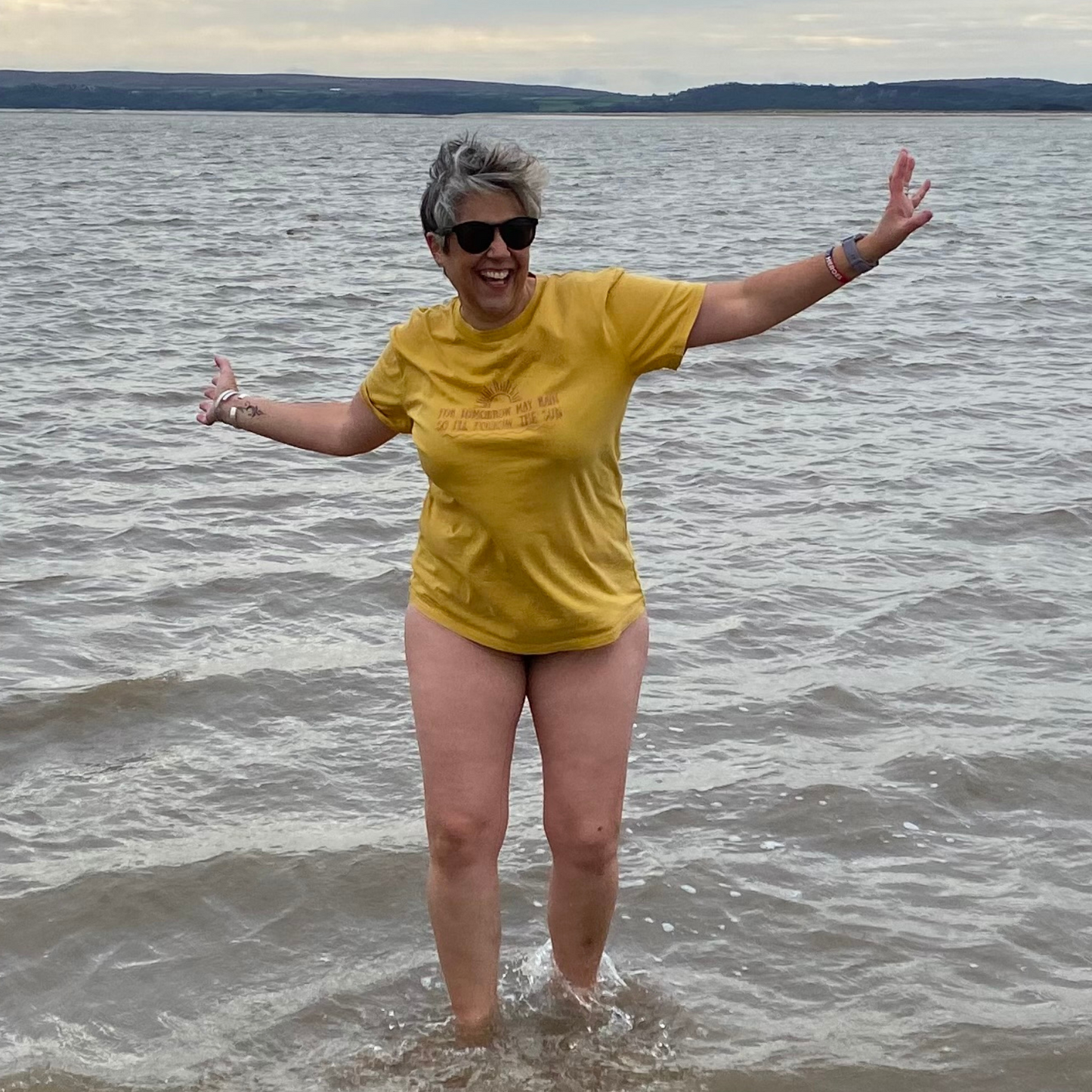 A woman dancing in the sea wearing a vintage gold t-shirt and sunglasses.