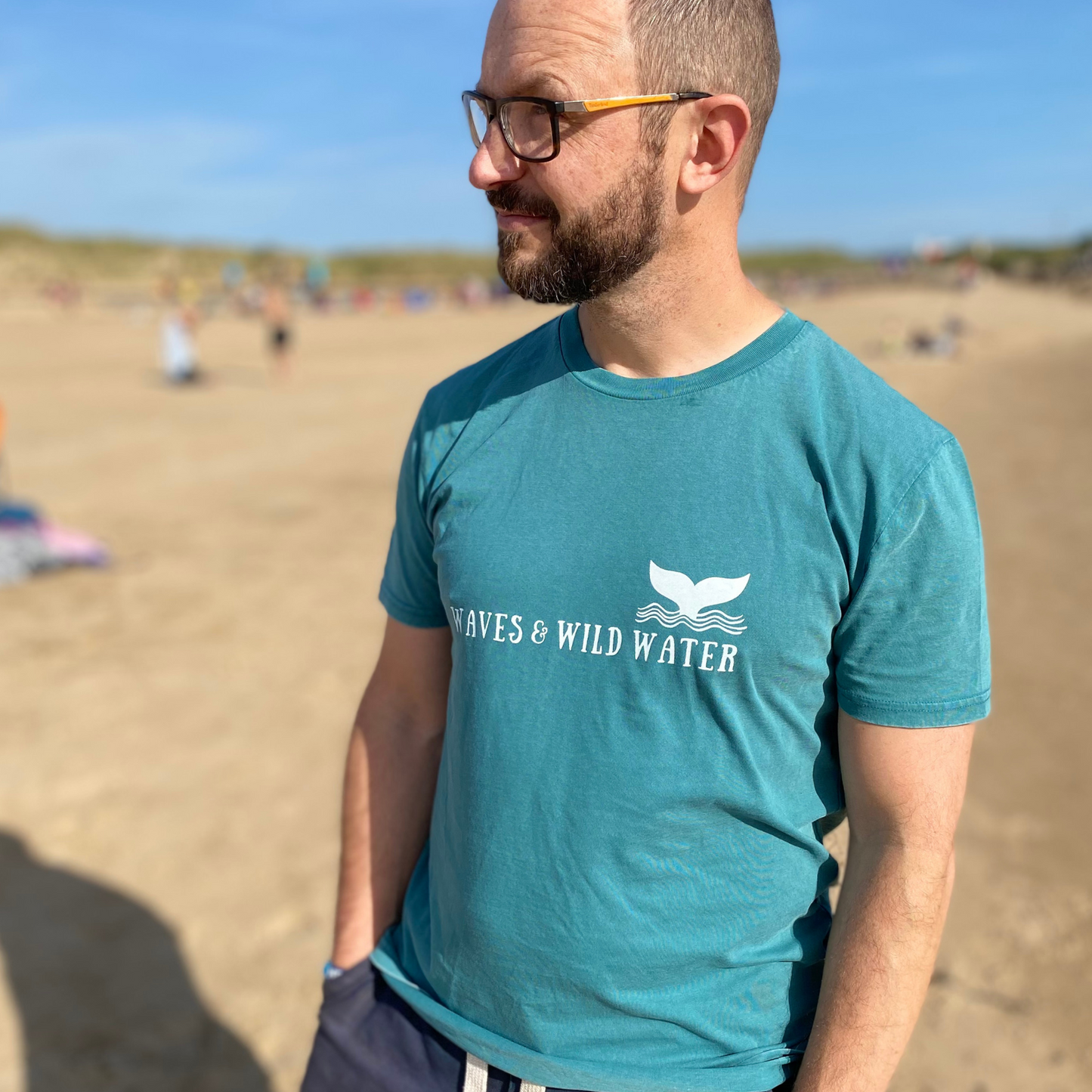 A man on a beach wearing glasses and a green Waves & Wild Water t-shirt.