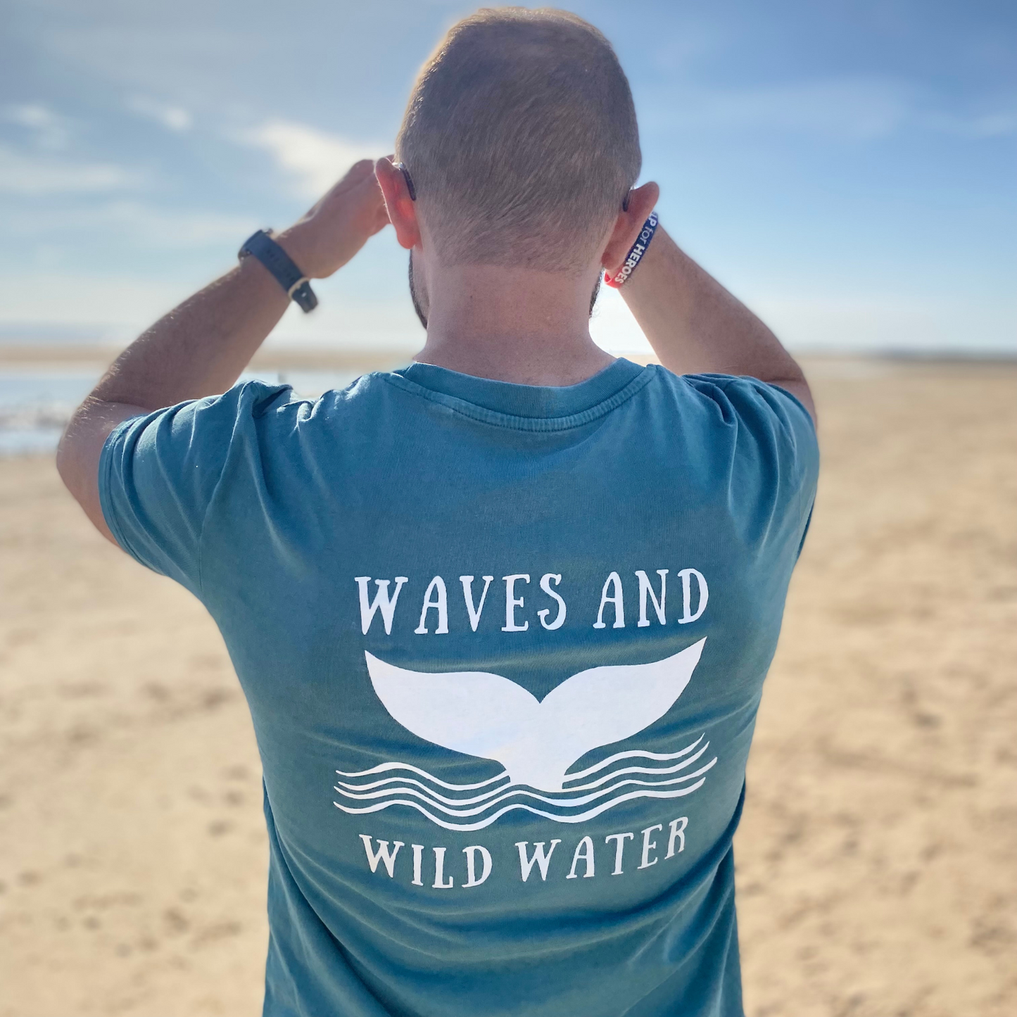 The back of a man stood on a beach, wearing a green Waves & Wild Water t-shirt.