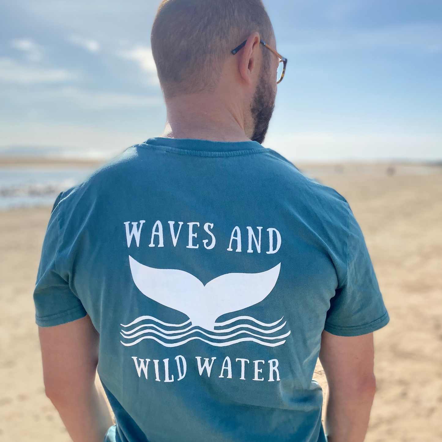 The back of a man wearing a green t-shirt with the Waves & Wild Water logo, depicting a whale tail coming out of waves, printed in white ink.