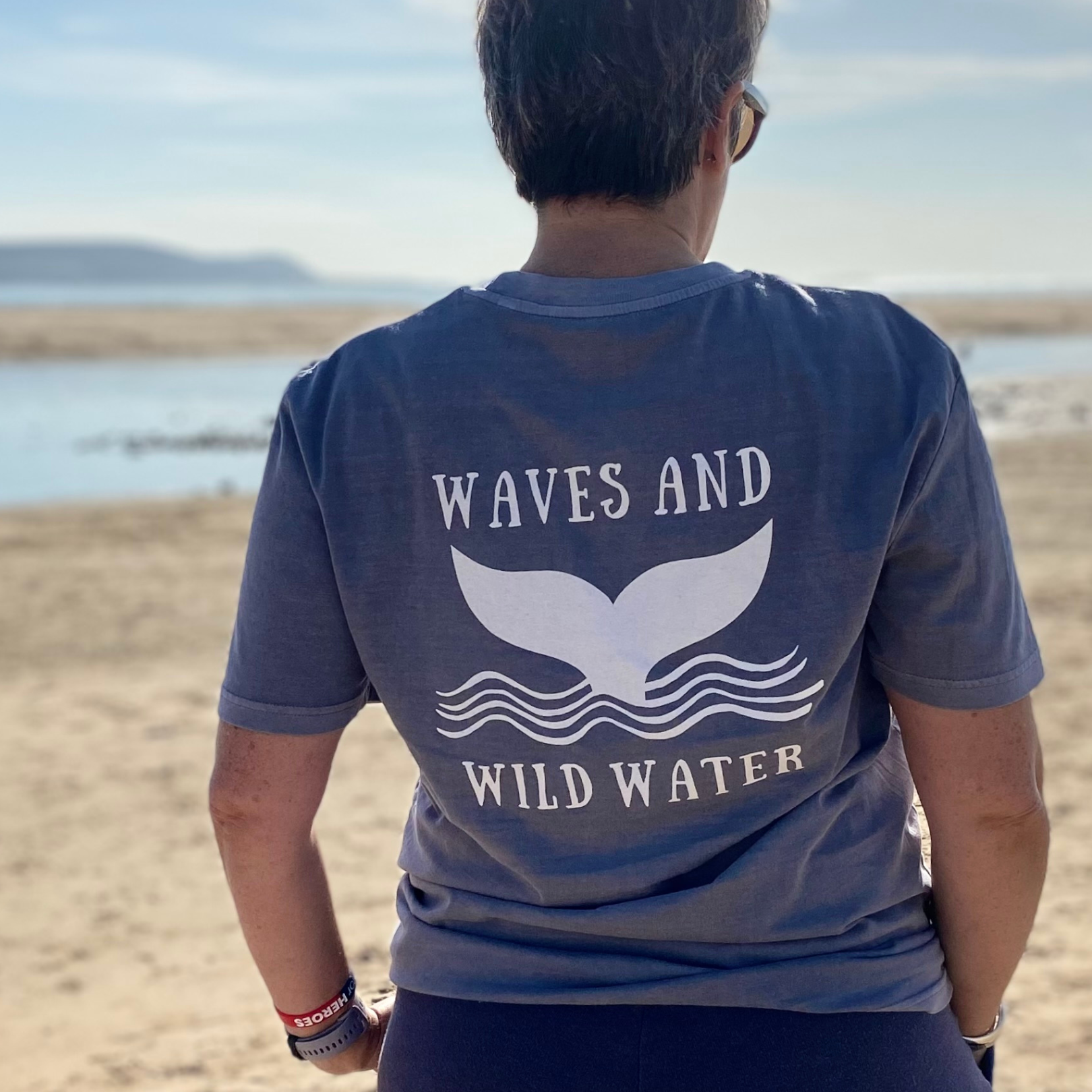 The back of a woman wearing a grey t-shirt with Waves & Wild Water logo, depicting a whale tail coming out of waves, printed in white ink.