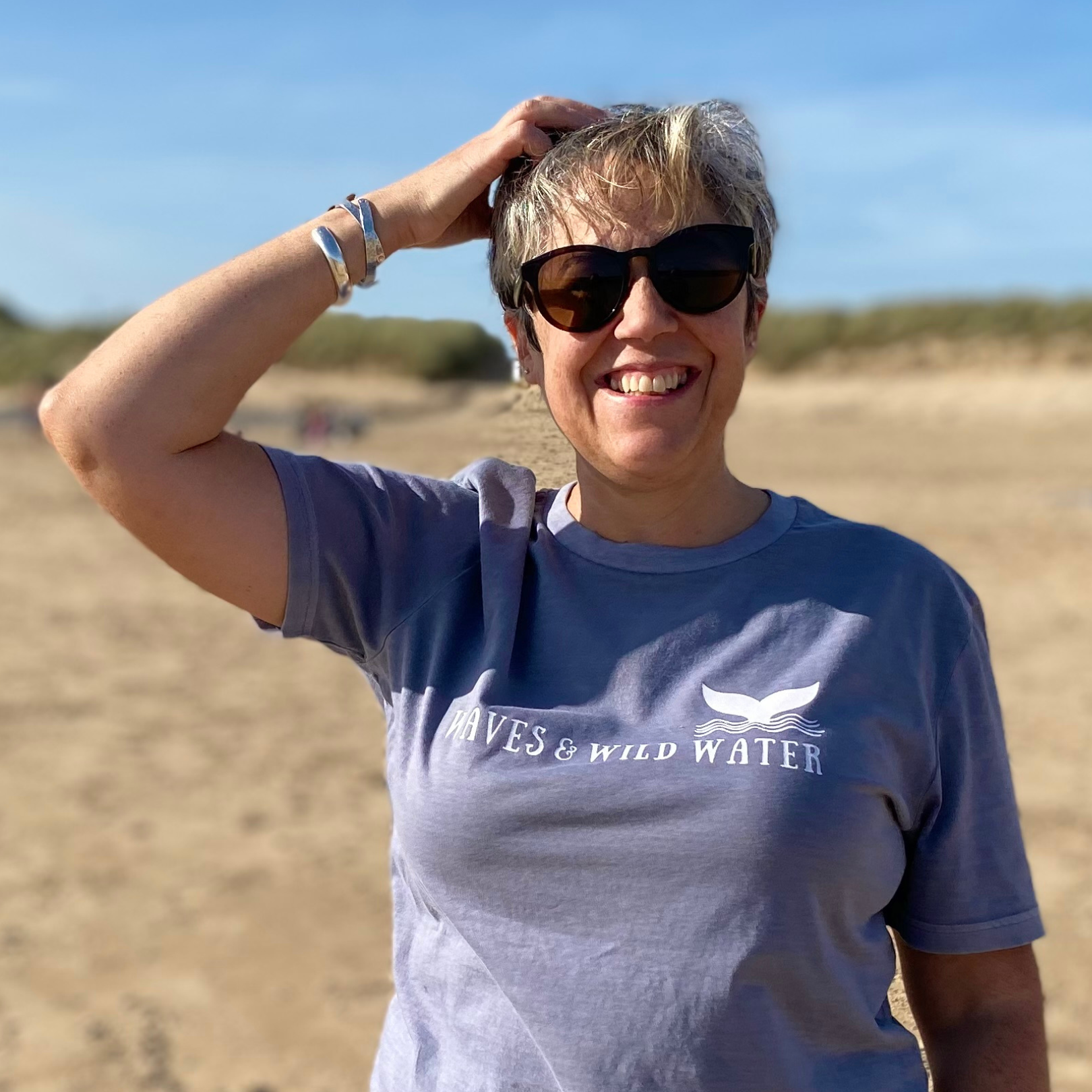 A woman on a beach wearing sunglasses and a grey Waves & Wild Water t-shirt.