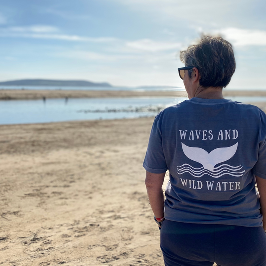 The back of a woman stood on a beach, wearing a grey Waves & Wild Water t-shirt.