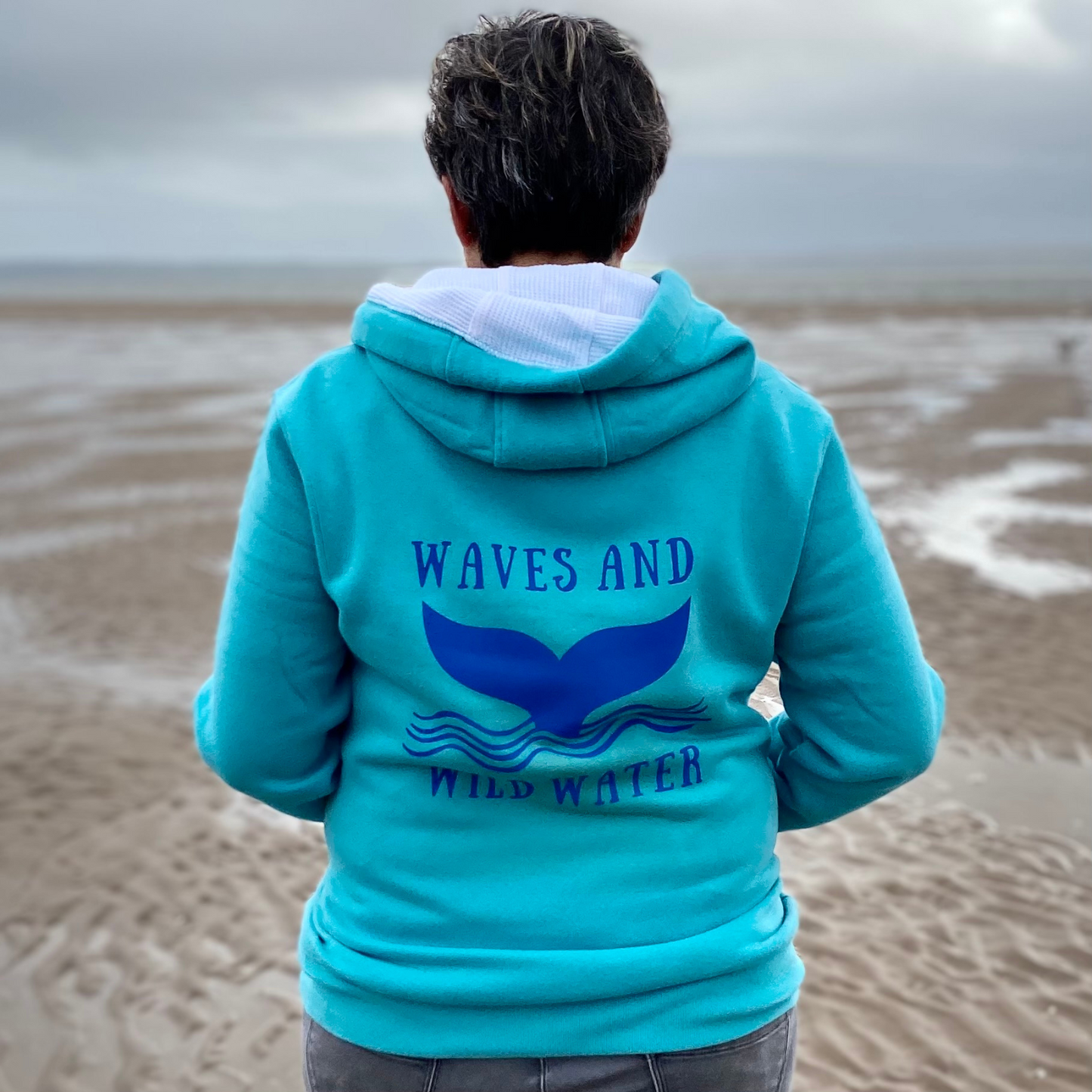 The back of a woman wearing a lagoon blue hoodie, with Waves & Wild Water logo printed on it in blue ink.