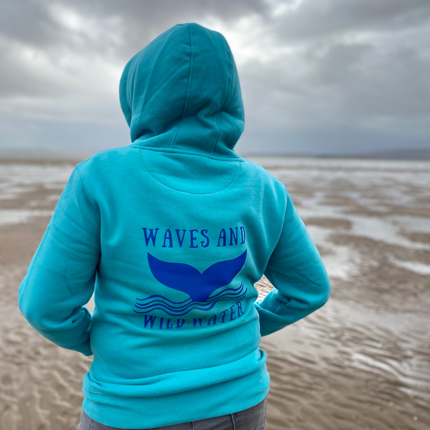 The back of a woman wearing a lagoon blue hoodie, with Waves & Wild Water logo printed on it in blue ink.