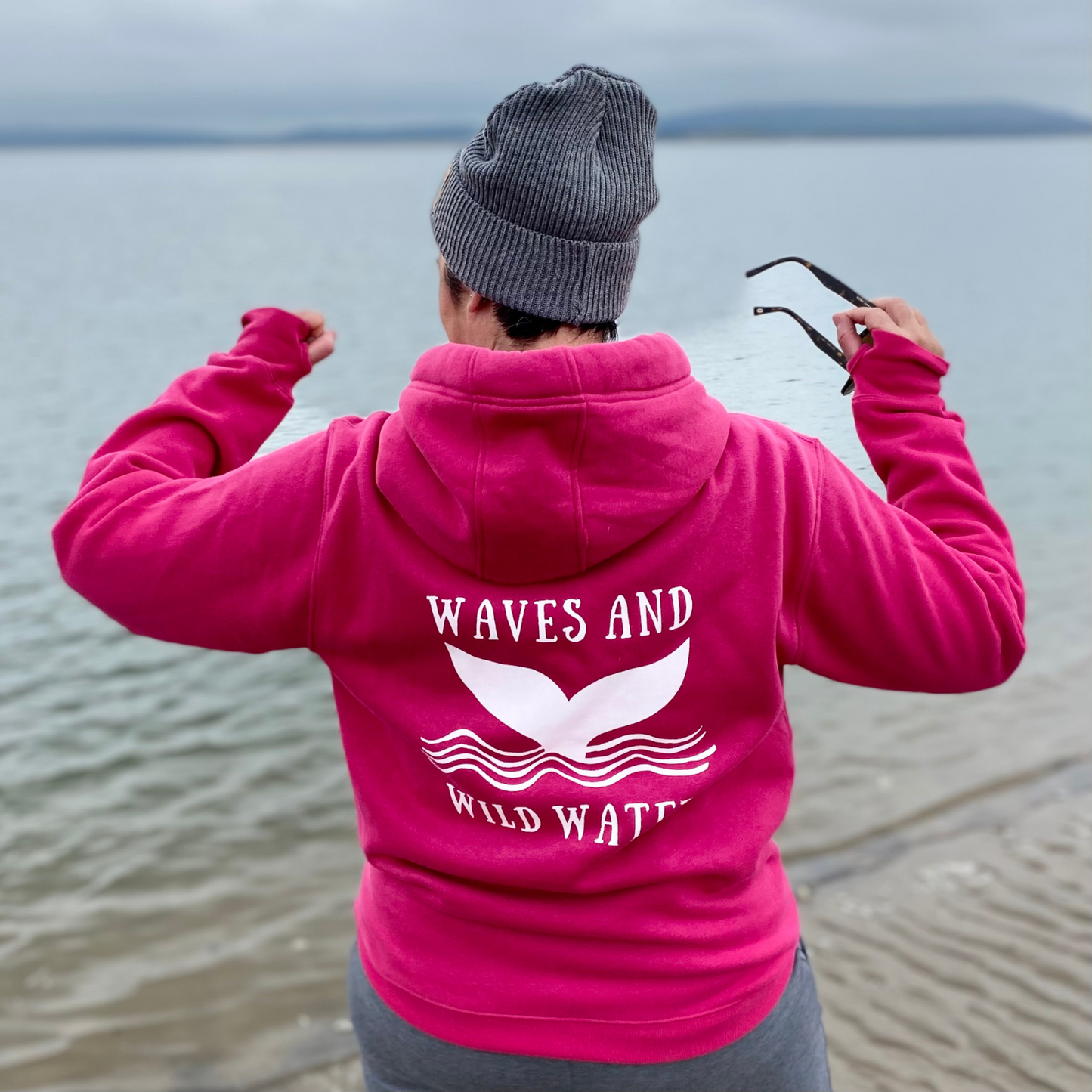 The back of a woman wearing a bright pink hoodie, with Waves & Wild Water logo printed on it in white ink.