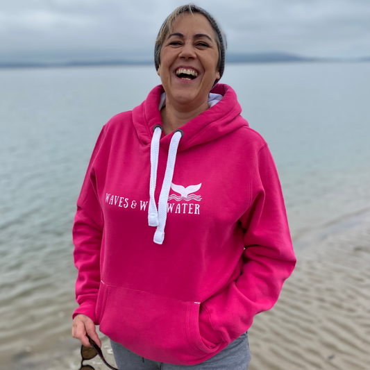 A woman wearing a bright pink hoodie with Waves & Wild Water printed across the front in white ink.