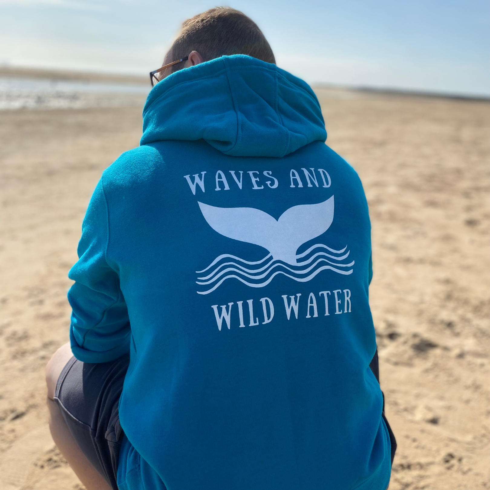 The back of a man wearing a tropical blue hoodie, with Waves & Wild Water logo printed on it in white ink.