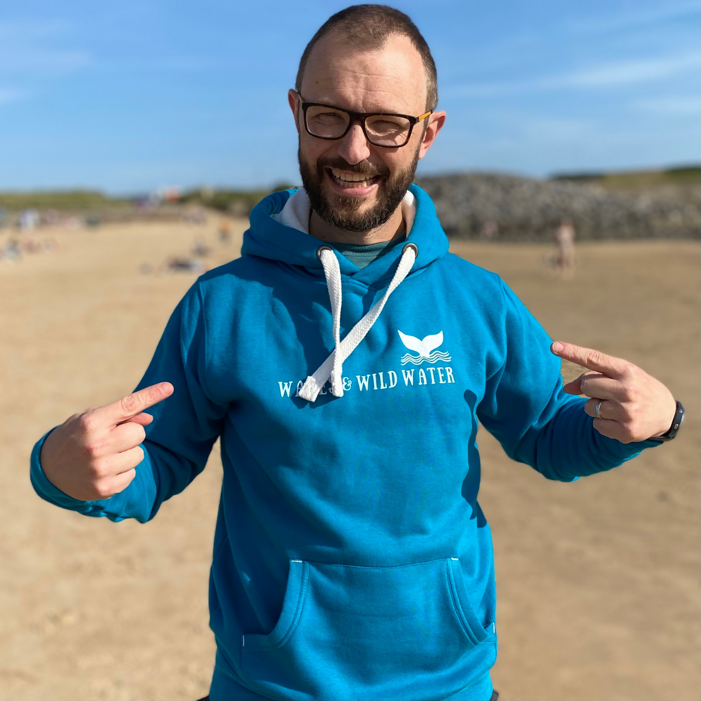 A man wearing a tropical blue hoodie and pointing to the Waves & Wild Water logo printed across his chest.